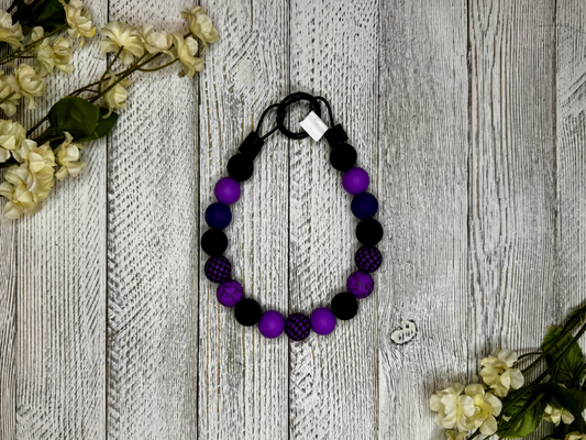 Beaded dog necklace with purple and black pattern including checkers. Displayed on a wooden surface with flowers for decoration.
