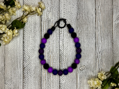 Beaded dog necklace with a purple and black pattern including checkers. Displayed on a wooden surface with flowers for decoration.