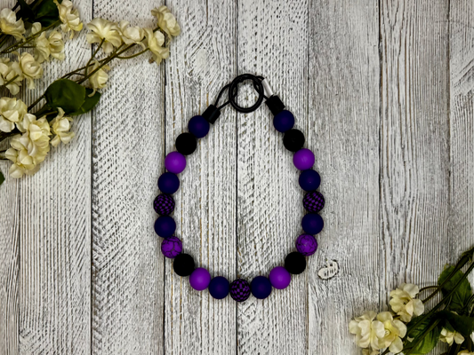 Beaded dog necklace with a purple and black pattern including checkers. Displayed on a wooden surface with flowers for decoration.