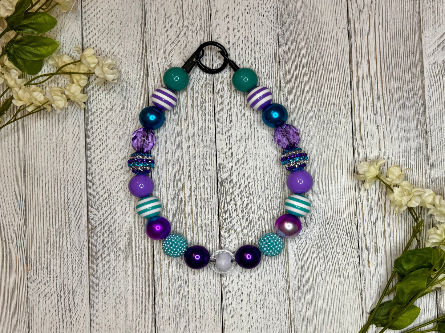 Beaded dog necklace with a purple and teal pattern including stripes. Displayed on a wooden surface with flowers for decoration.