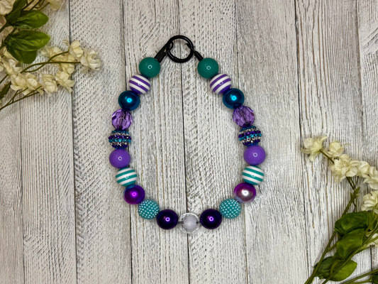 Beaded dog necklace with a purple and teal pattern including stripes. Displayed on a wooden surface with flowers for decoration.