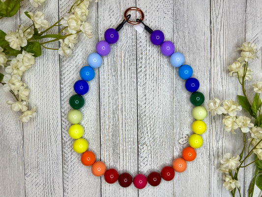Beaded dog necklace with a rainbow pattern. Displayed on a wooden surface with flowers for decoration.