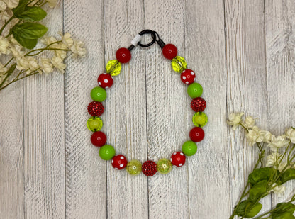 Beaded dog necklace with a red and green pattern including polka dots. Displayed on a wooden surface with flowers for decoration.