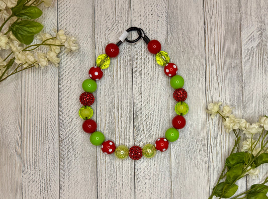 Beaded dog necklace with a red and green pattern including polka dots. Displayed on a wooden surface with flowers for decoration.