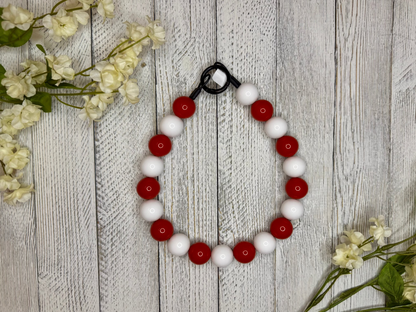 Beaded dog necklace with a red and white pattern. Displayed on a wooden surface with flowers for decoration.