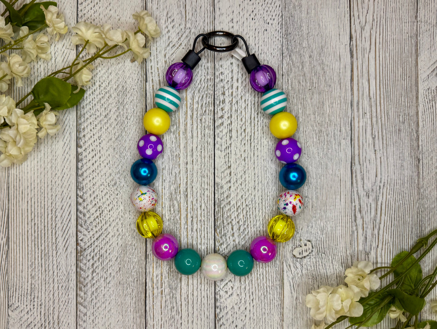 Beaded dog necklace with a retro pattern including purples, teals, and yellows. Displayed on a wooden surface with flowers for decoration.