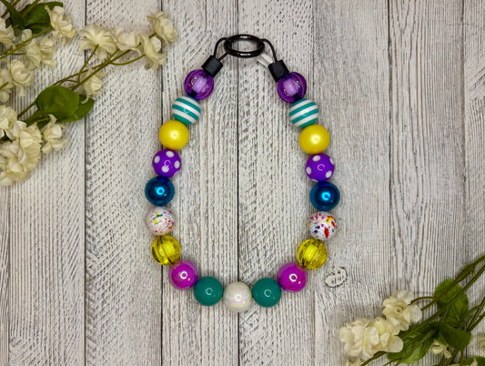 Beaded dog necklace with a retro pattern including purples, teals, and yellows. Displayed on a wooden surface with flowers for decoration.