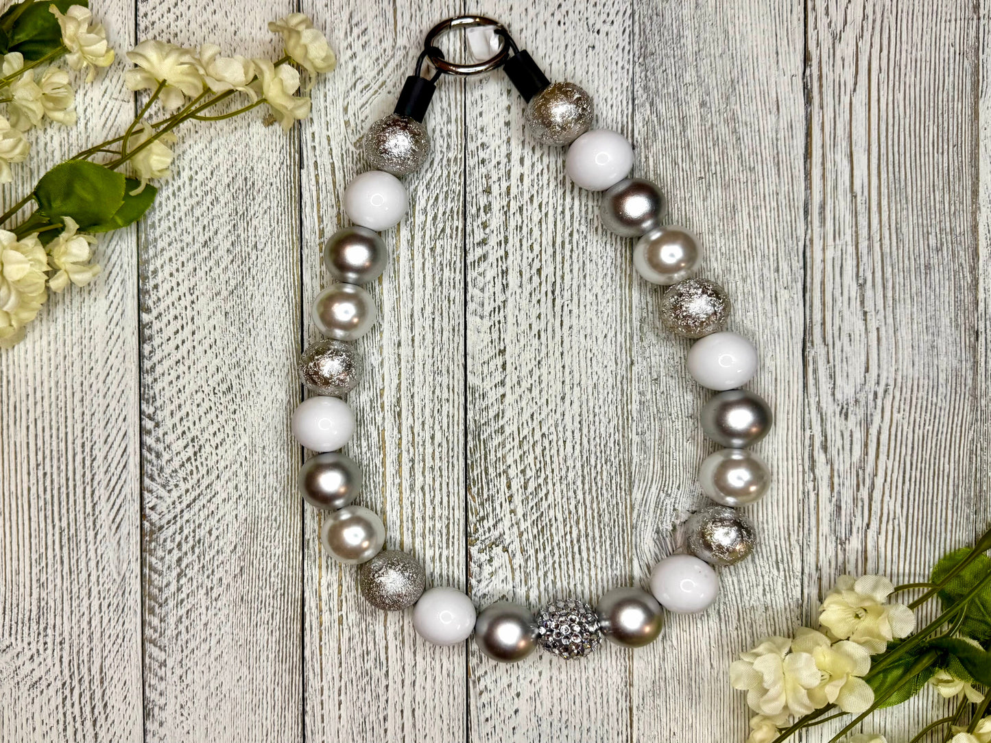 Beaded pet necklace with a silver and white pattern, including sparkles. Displayed on a wooden surface with flowers for decoration. 