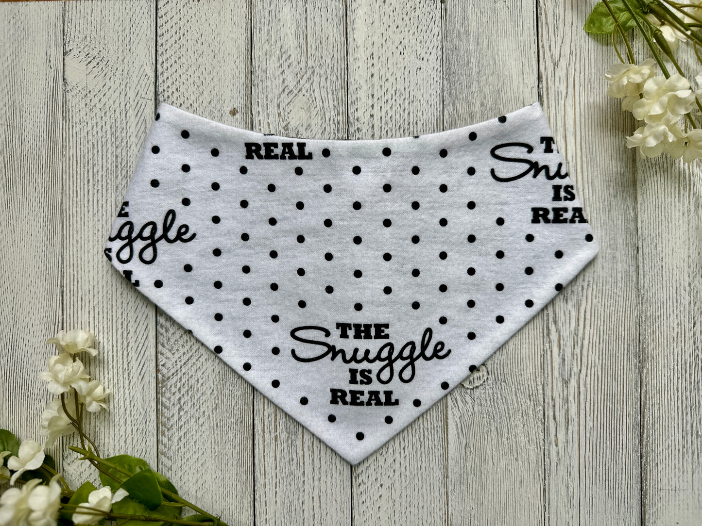 A pet bandana with a white background featuring a polka dot and snuggle is real pattern, displayed on a wooden surface with flowers for decoration.