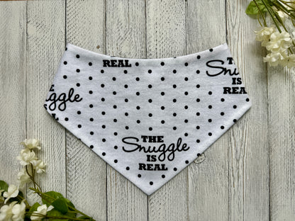 A pet bandana with a white background featuring a polka dot and snuggle is real pattern, displayed on a wooden surface with flowers for decoration.