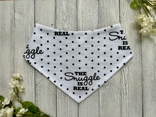 A pet bandana with a white background featuring a polka dot and snuggle is real pattern, displayed on a wooden surface with flowers for decoration.