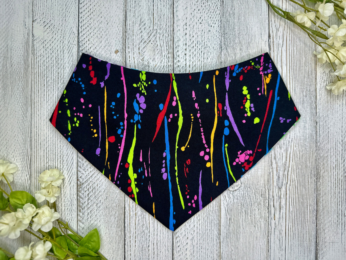 A pet bandana with a black background featuring rainbow splatter design, displayed on a wooden surface with flowers for decoration. 