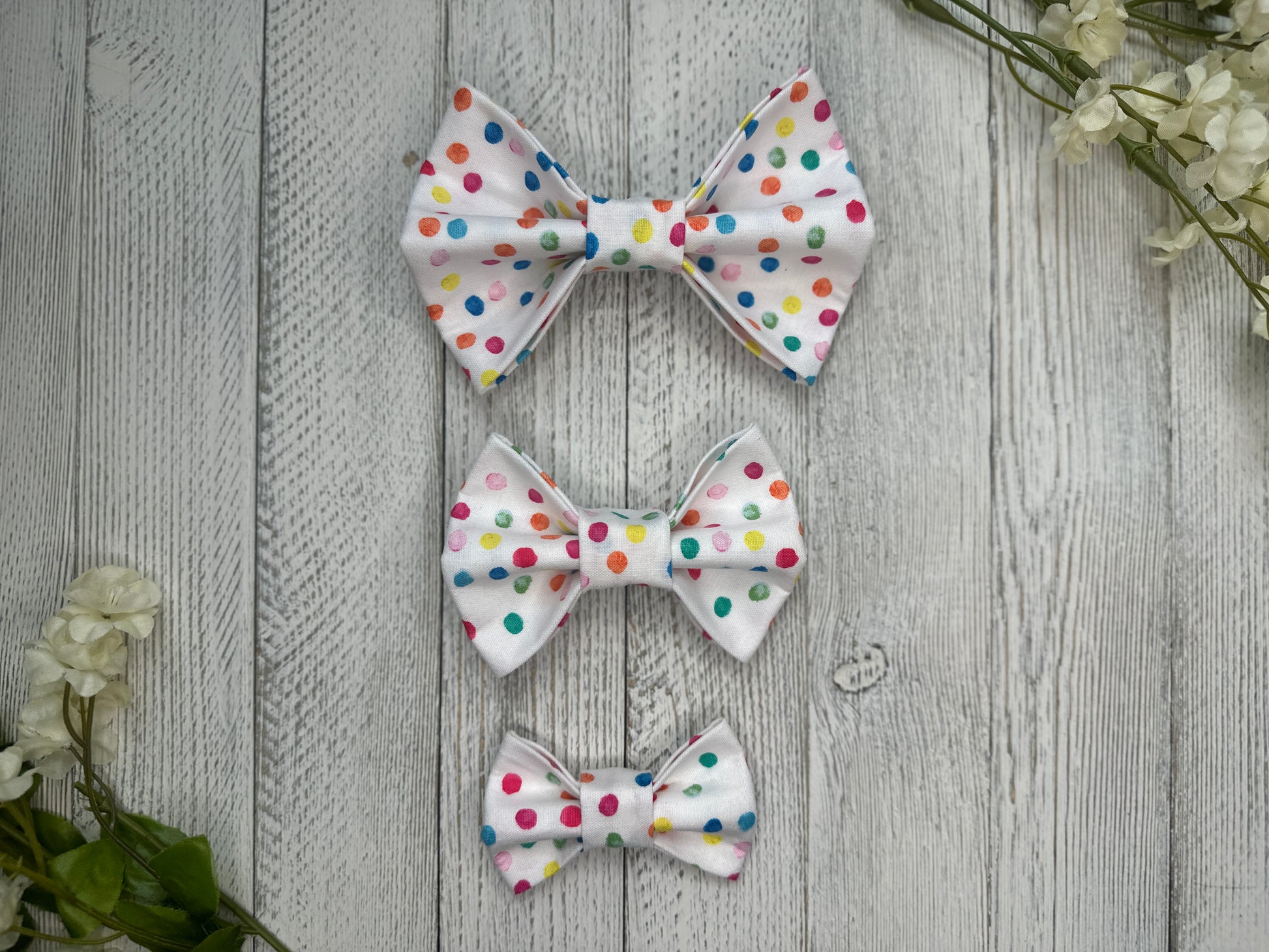 Three pet bow ties (small, medium, large) with a white background and rainbow polka dot pattern, displayed on a wooden surface with flowers for decoration.