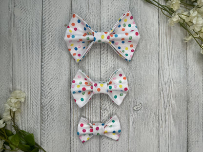 Three pet bow ties (small, medium, large) with a white background and rainbow polka dot pattern, displayed on a wooden surface with flowers for decoration.
