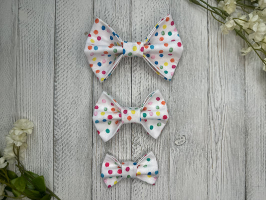 Three pet bow ties (small, medium, large) with a white background and rainbow polka dot pattern, displayed on a wooden surface with flowers for decoration.