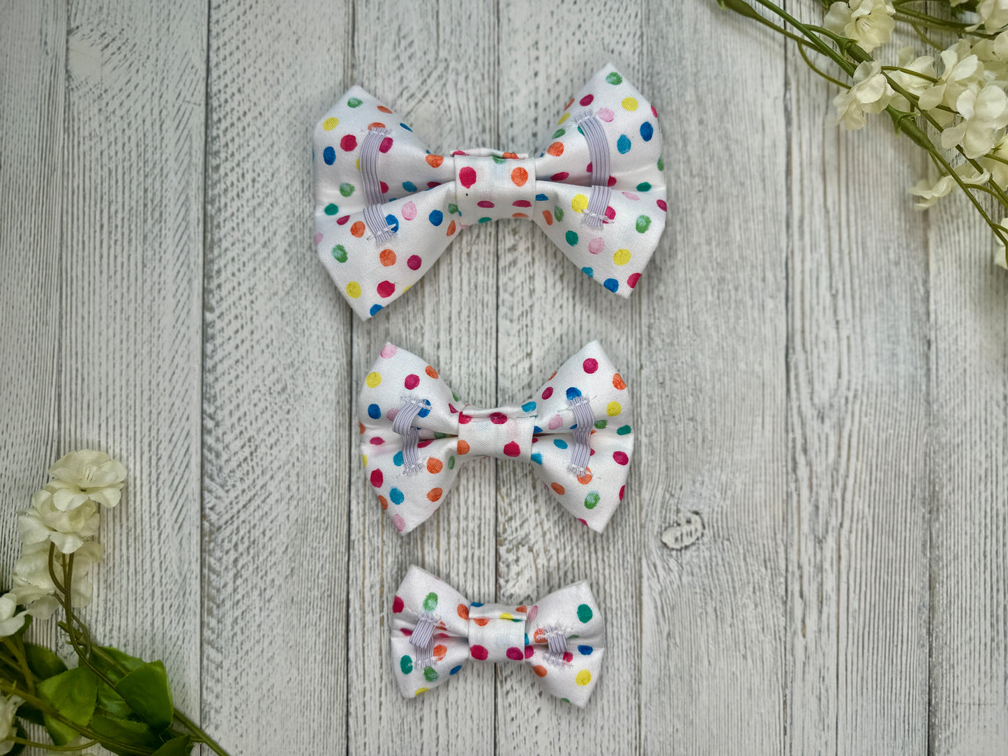 Picture of the backside of pet bow ties showing the elastic straps that fit around a collar. Displayed on a wooden surface with flowers for decoration. 
