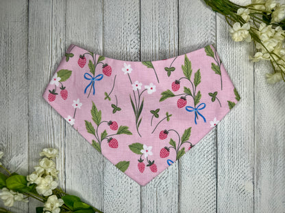 A pet bandana with a pink background featuring a strawberry pattern with white flowers, displayed on a wooden surface with flowers for decoration. 