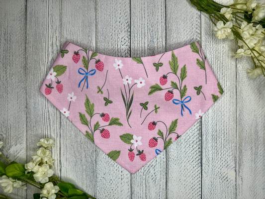 A pet bandana with a pink background featuring a strawberry pattern with white flowers, displayed on a wooden surface with flowers for decoration. 