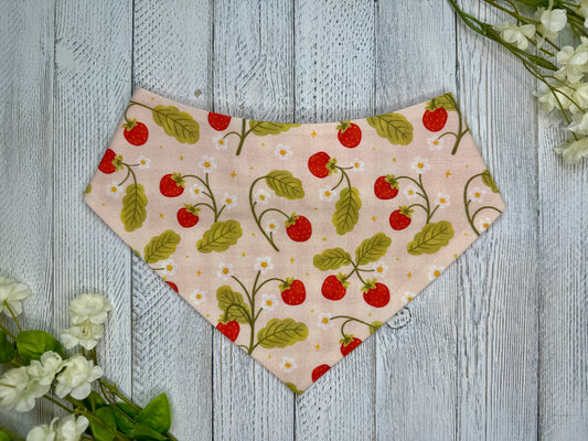 A pet bandana with a light pink background featuring a strawberry pattern with white flowers, displayed on a wooden surface with flowers for decoration. 