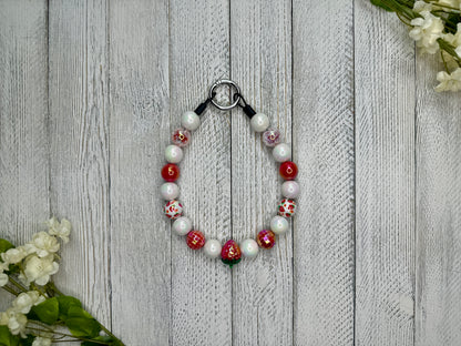Beaded pet necklace with a red and white pattern including strawberries. Displayed on a wooden surface with flowers for decoration. 