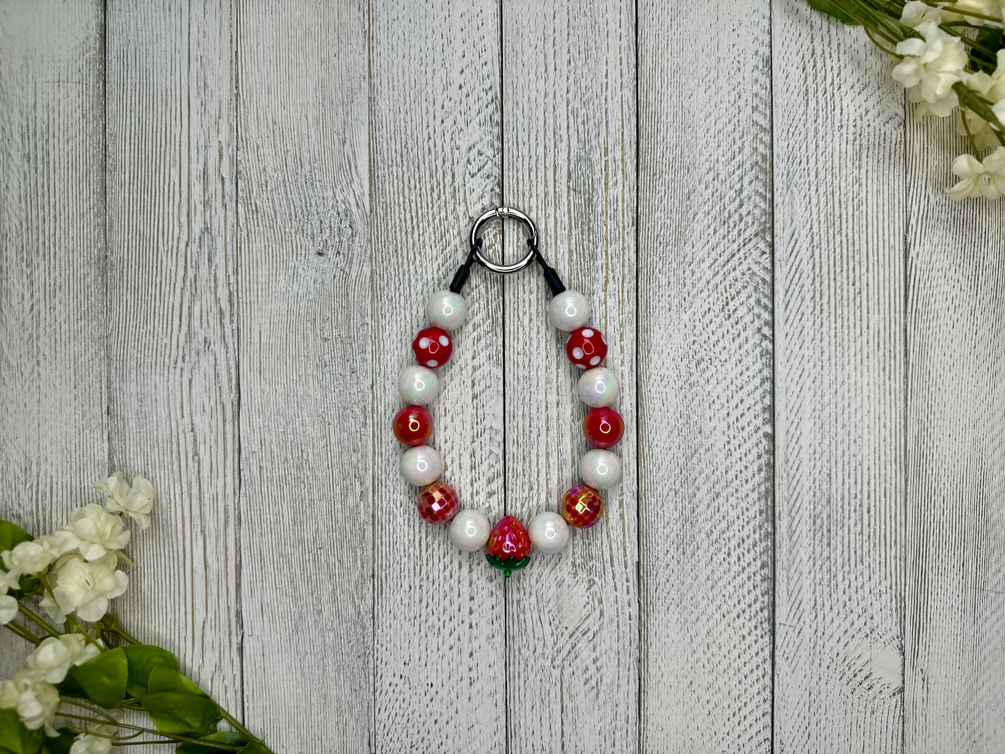 Beaded pet necklace with a red and white pattern including polka dots and strawberries. Displayed on a wooden surface with flowers for decoration. 