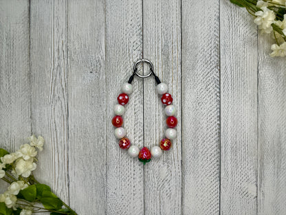 Beaded pet necklace with a red and white pattern including polka dots and strawberries. Displayed on a wooden surface with flowers for decoration. 
