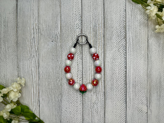 Beaded pet necklace with a red and white pattern including polka dots and strawberries. Displayed on a wooden surface with flowers for decoration. 