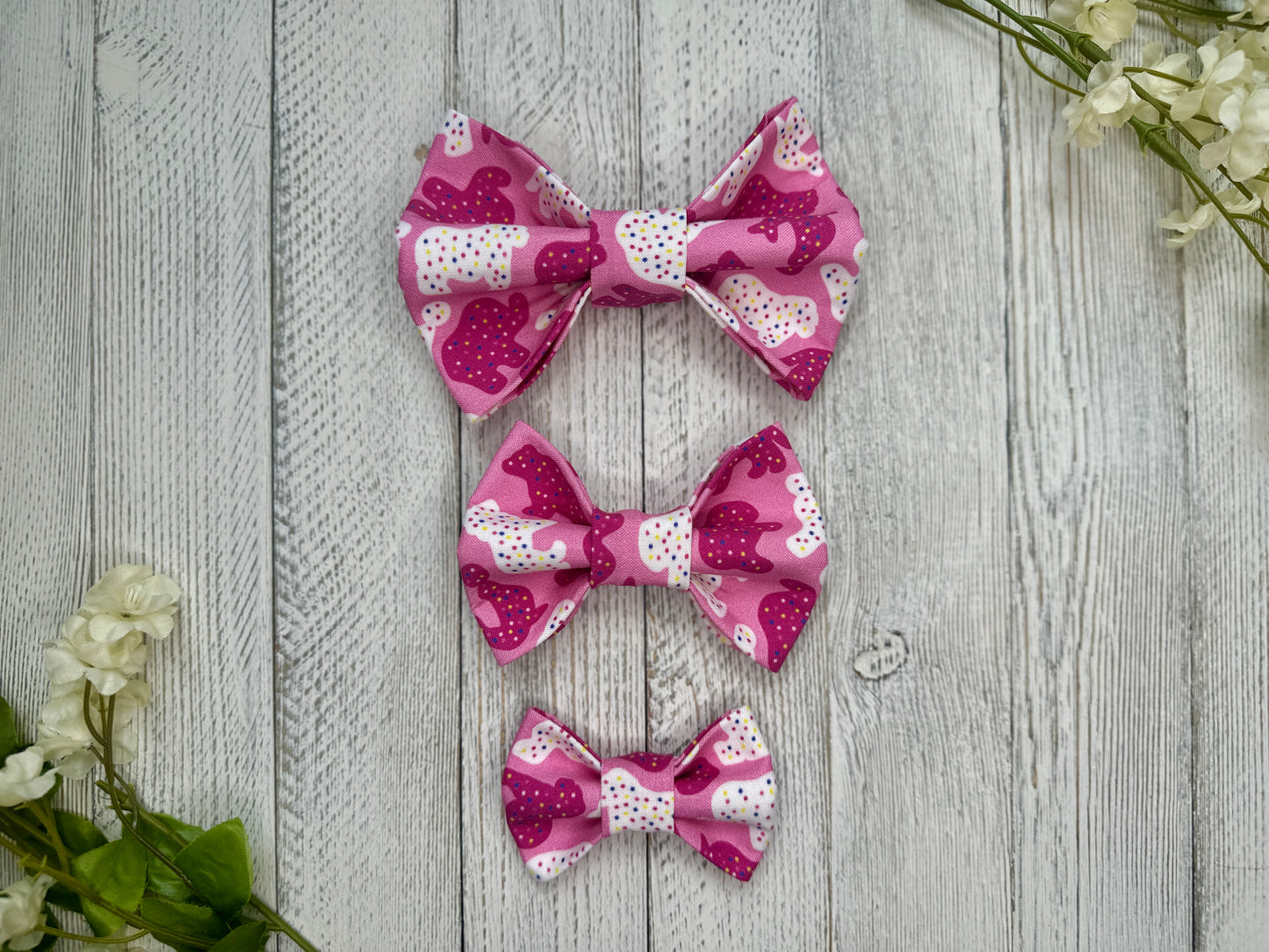 Three pet bow ties (small, medium, large) with a pink background and animal cracker pattern, displayed on a wooden surface with flowers for decoration. 