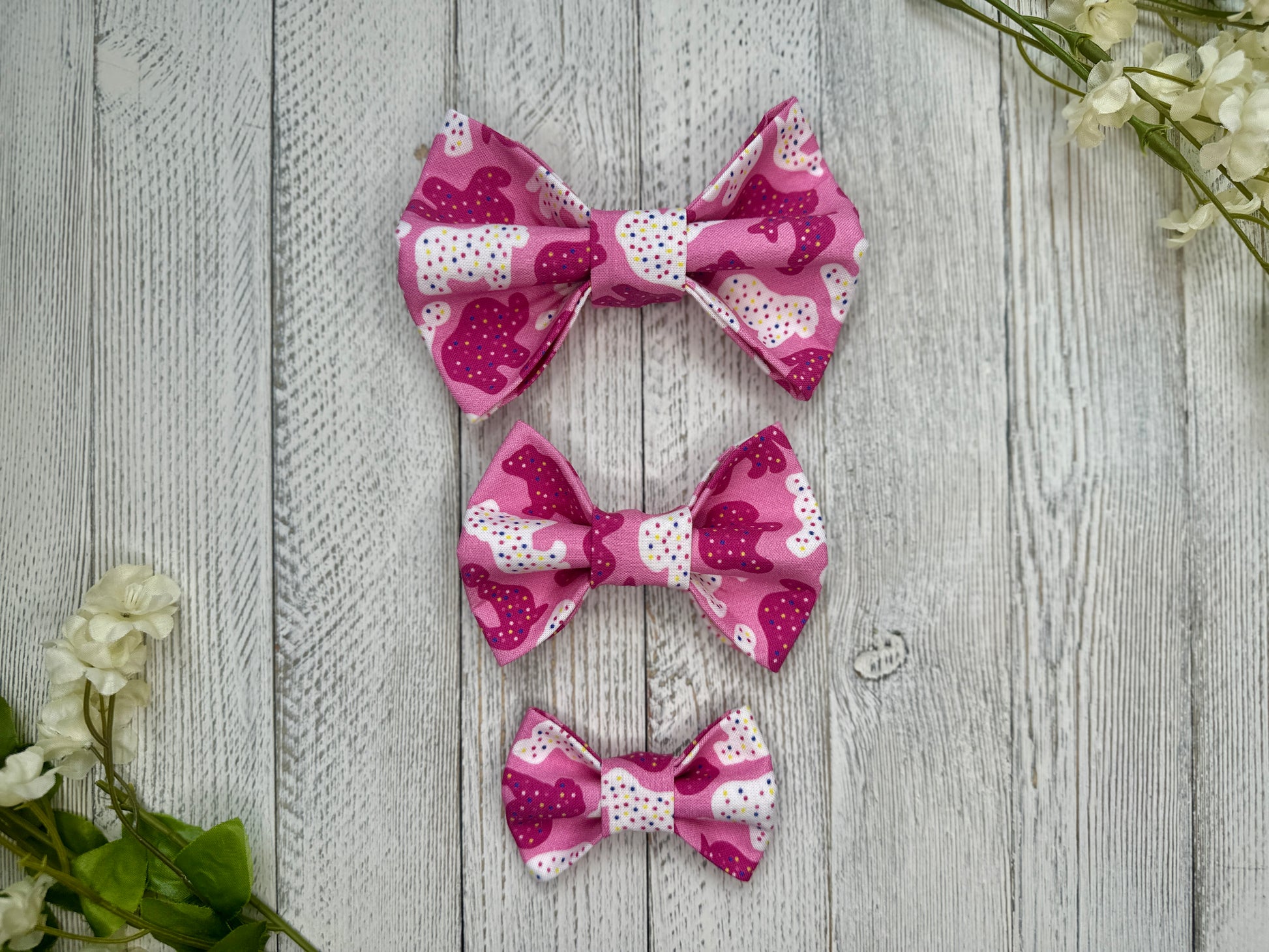 Three pet bow ties (small, medium, large) with a pink background and animal cracker pattern, displayed on a wooden surface with flowers for decoration. 