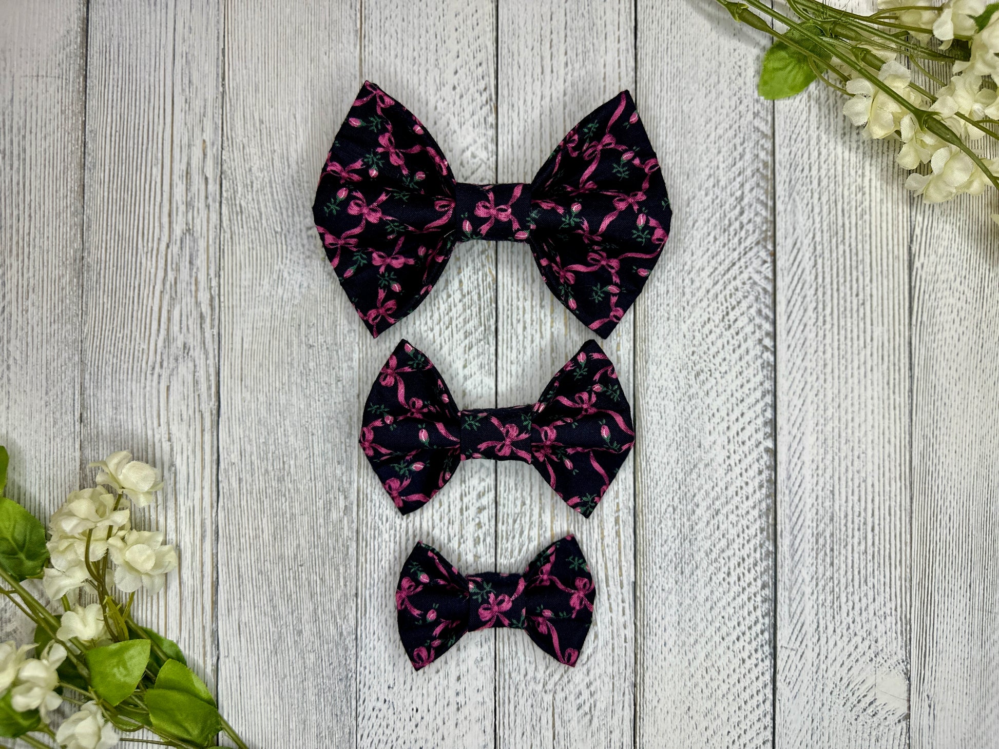 Three pet bow ties (small, medium, large) with a black background and rose and pink bow pattern displayed on a wooden surface with flowers for decoration. 
