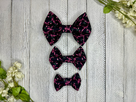 Three pet bow ties (small, medium, large) with a black background and rose and pink bow pattern displayed on a wooden surface with flowers for decoration. 