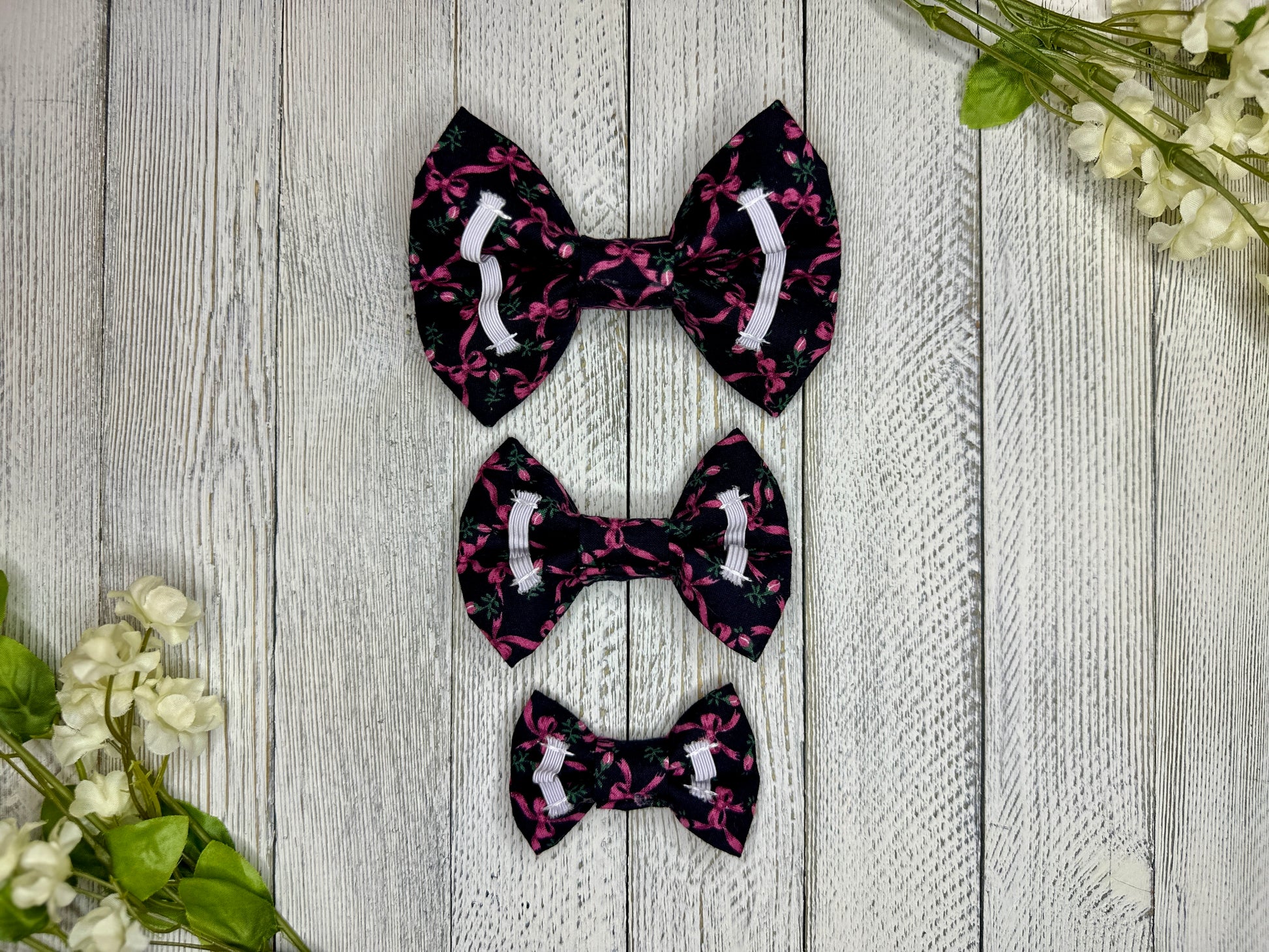 Picture of the backside of pet bow ties showing the elastic straps that fit around a collar. Displayed on a wooden surface with flowers for decoration. 