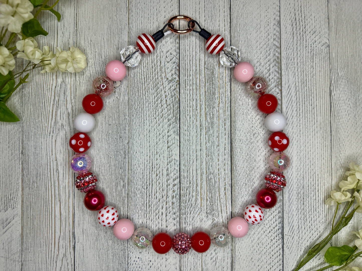 Beaded pet necklace with a red, pink, and white pattern, including hearts. Displayed on a wooden surface with flowers for decoration. 