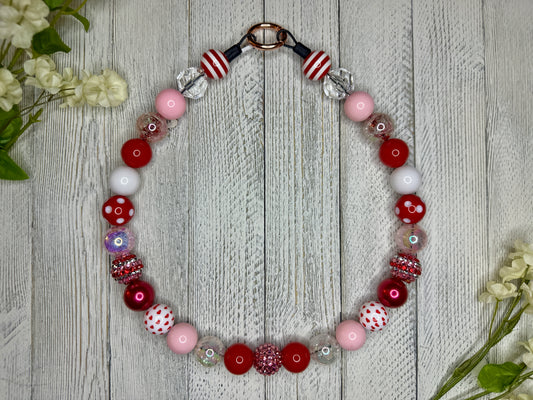 Beaded pet necklace with a red, pink, and white pattern, including hearts. Displayed on a wooden surface with flowers for decoration. 