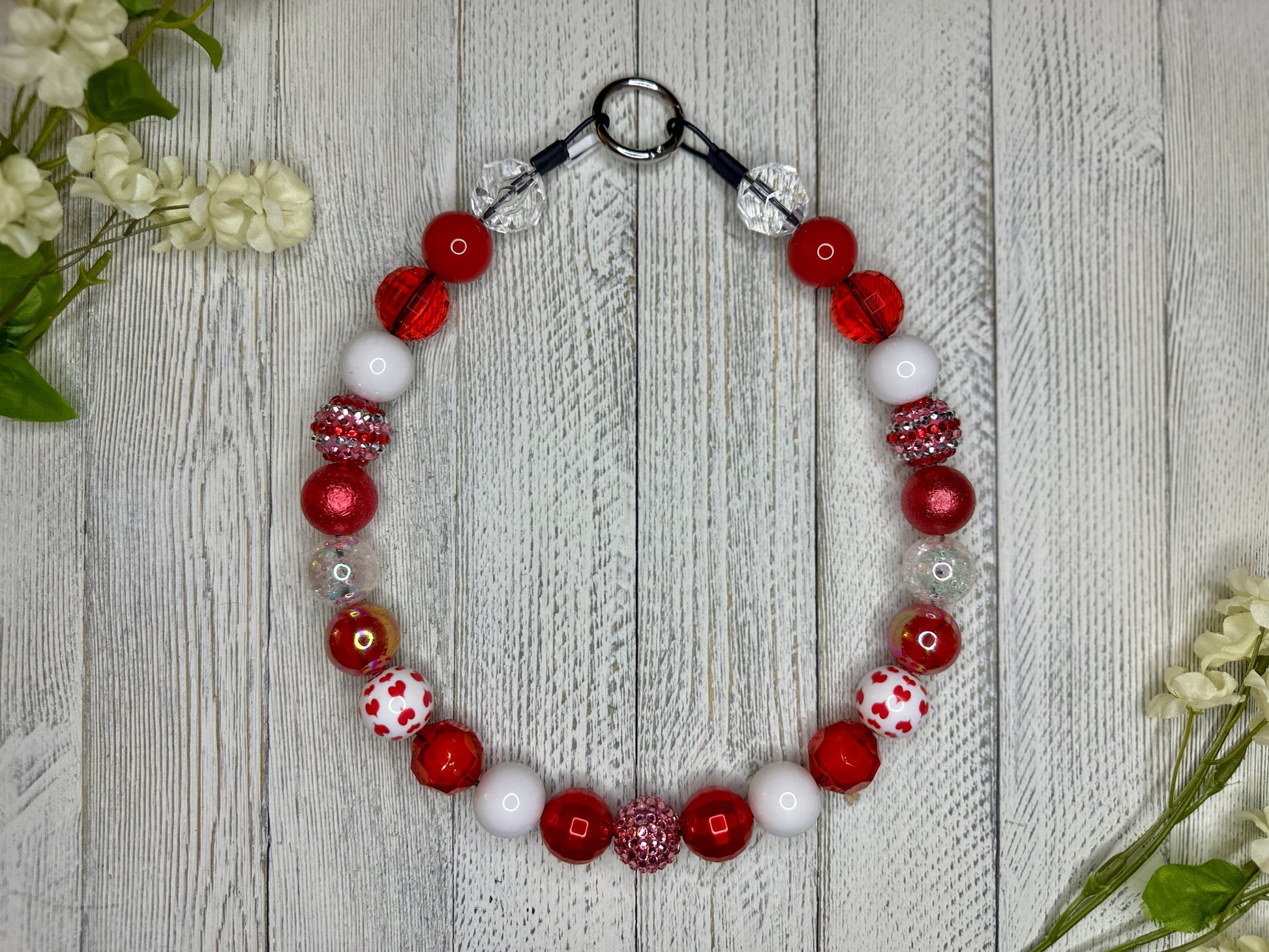 Beaded dog necklace with a red and white pattern including hearts. Displayed on a wooden surface with flowers for decoration. 