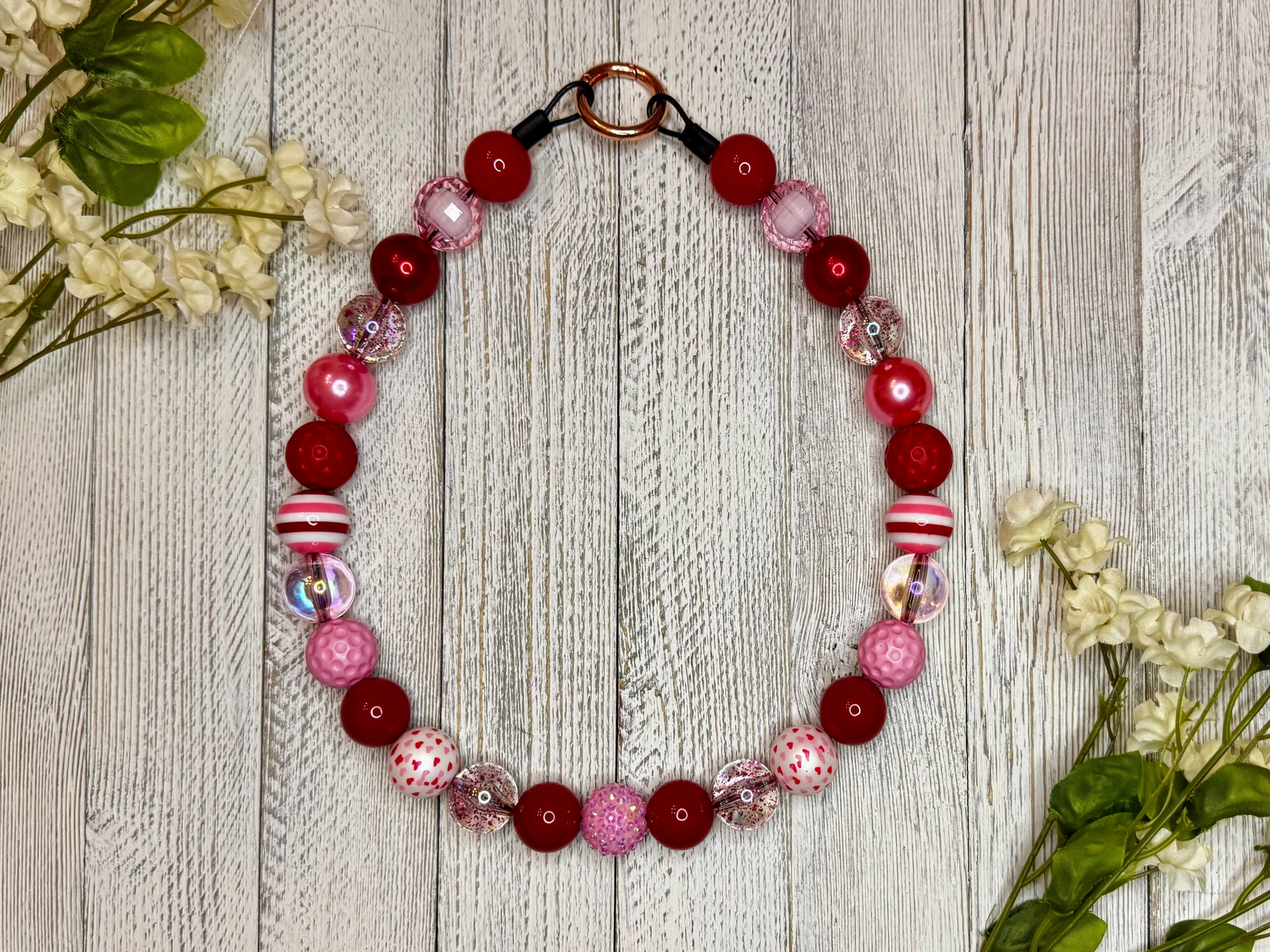 Beaded dog necklace with a red, pink, and white pattern including hearts and stripes. Displayed on a wooden surface with flowers for decoration.
