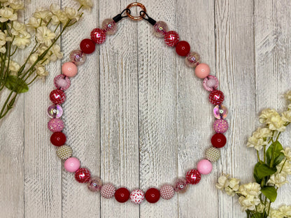 Beaded dog necklace with a red, pink and white pattern including sprinkles nad zig zags. Displayed on a wooden surface with flowers for decoration.
