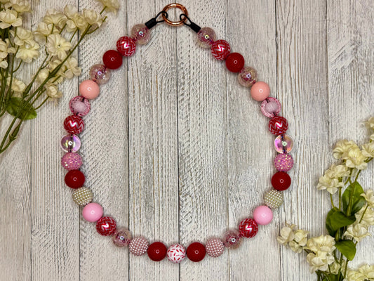 Beaded dog necklace with a red, pink and white pattern including sprinkles nad zig zags. Displayed on a wooden surface with flowers for decoration.