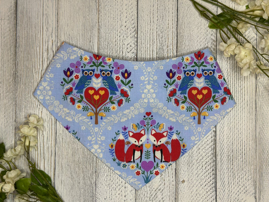A pet bandana with a light blue background featuring a fox and owl pattern with flowers and hearts, displayed on a wooden surface with flowers for decoration. 
