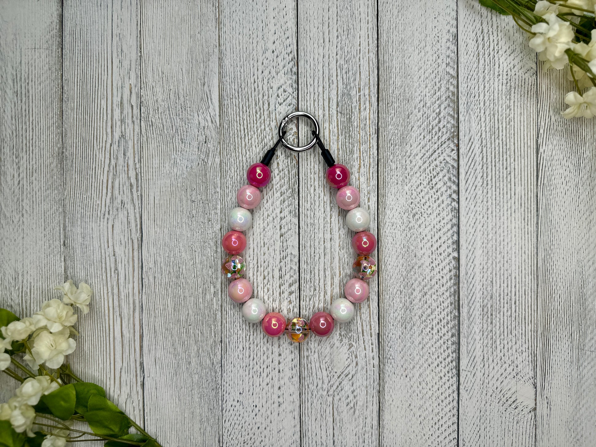 Beaded pet necklace with a pink and white pattern including strawberries. Displayed on a wooden surface with flowers for decoration. 
