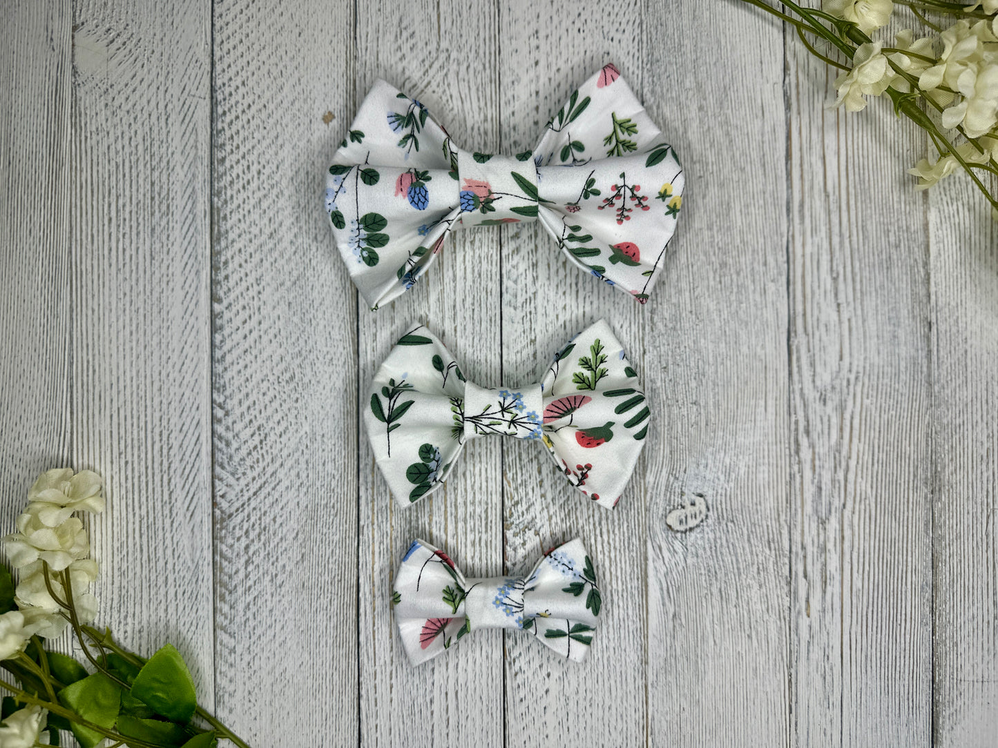 Three pet bow ties (small, medium, large) with a white background and wildflower pattern displayed on a wooden surface with flowers for decoration. 