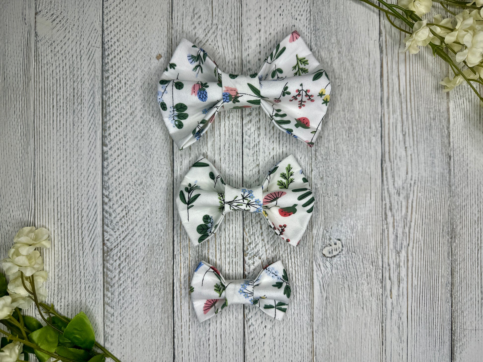 Three pet bow ties (small, medium, large) with a white background and wildflower pattern displayed on a wooden surface with flowers for decoration. 