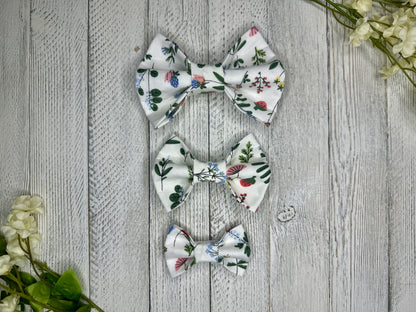 Three pet bow ties (small, medium, large) with a white background and wildflower pattern displayed on a wooden surface with flowers for decoration. 