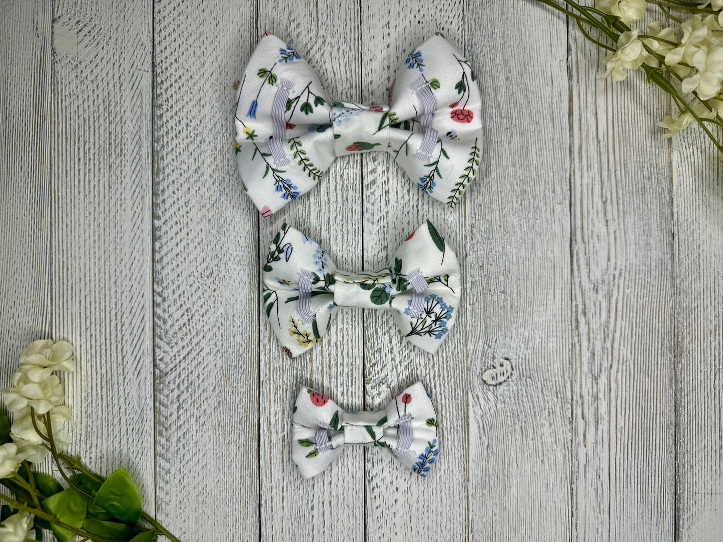Picture of the backside of pet bow ties showing the elastic straps that fit around a collar. Displayed on a wooden surface with flowers for decoration. 