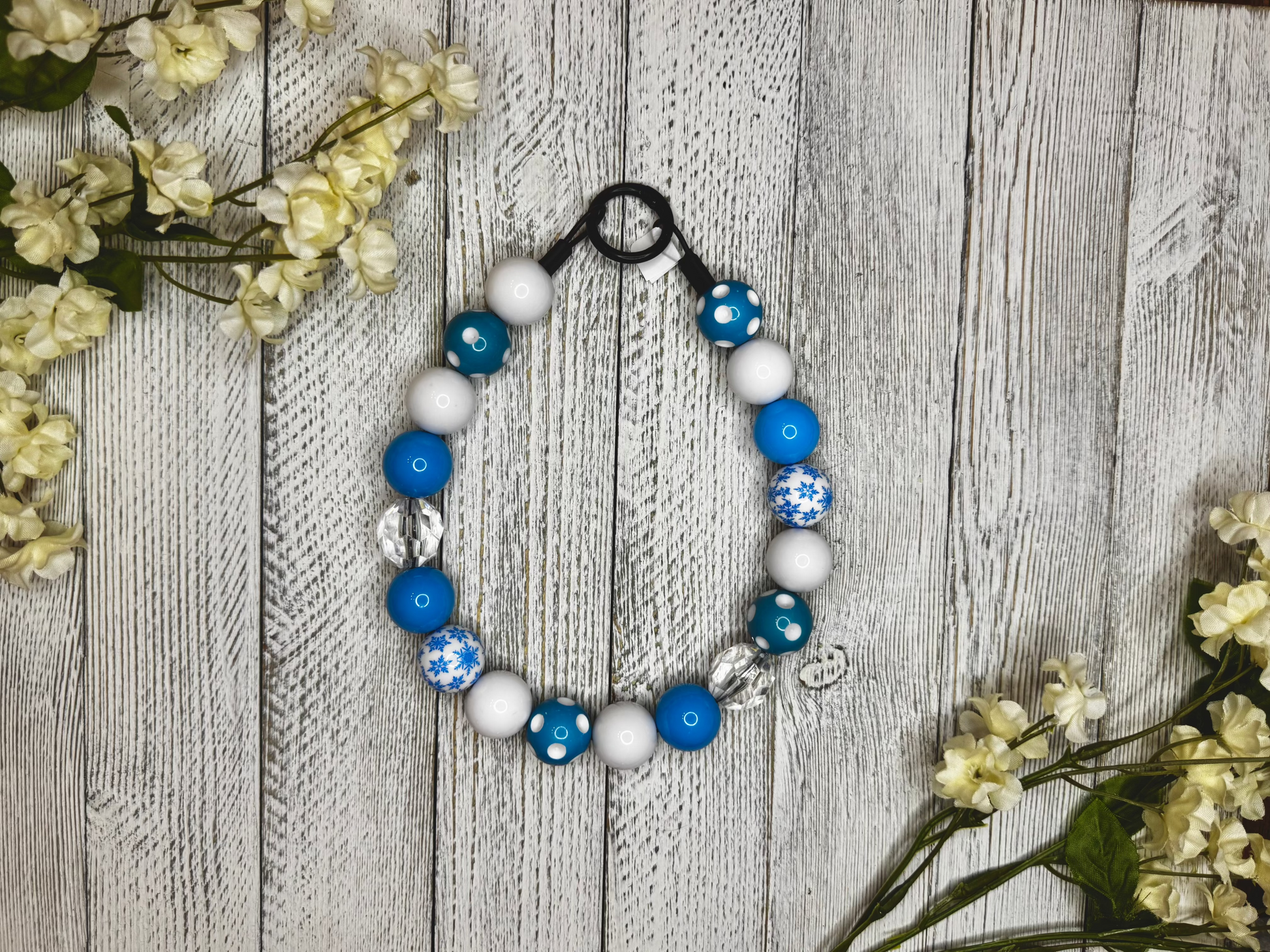Beaded dog necklace with a light blue and white pattern including snow flakes and polka dots. Displayed on a wooden surface with flowers for decoration.