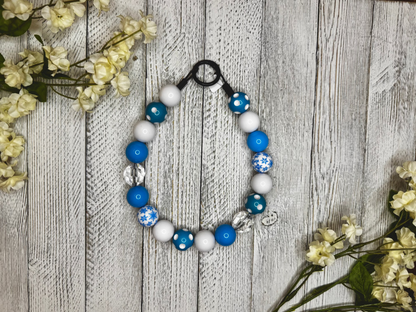 Beaded dog necklace with a light blue and white pattern including snow flakes and polka dots. Displayed on a wooden surface with flowers for decoration.