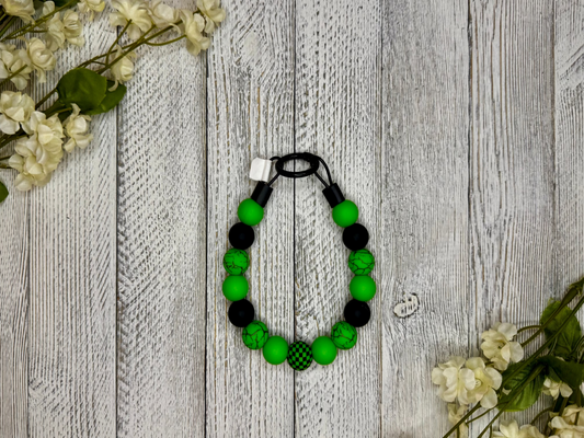 Beaded pet necklace with a green and black pattern, including checkers. Displayed on a wooden surface with flowers for decoration. 