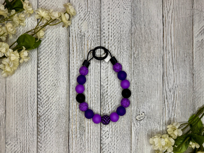 Beaded pet necklace with a purple and black pattern, including checkers. Displayed on a wooden surface with flowers for decoration.