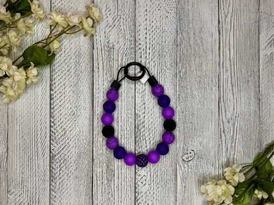 Beaded pet necklace with a purple and black pattern, including checkers. Displayed on a wooden surface with flowers for decoration.