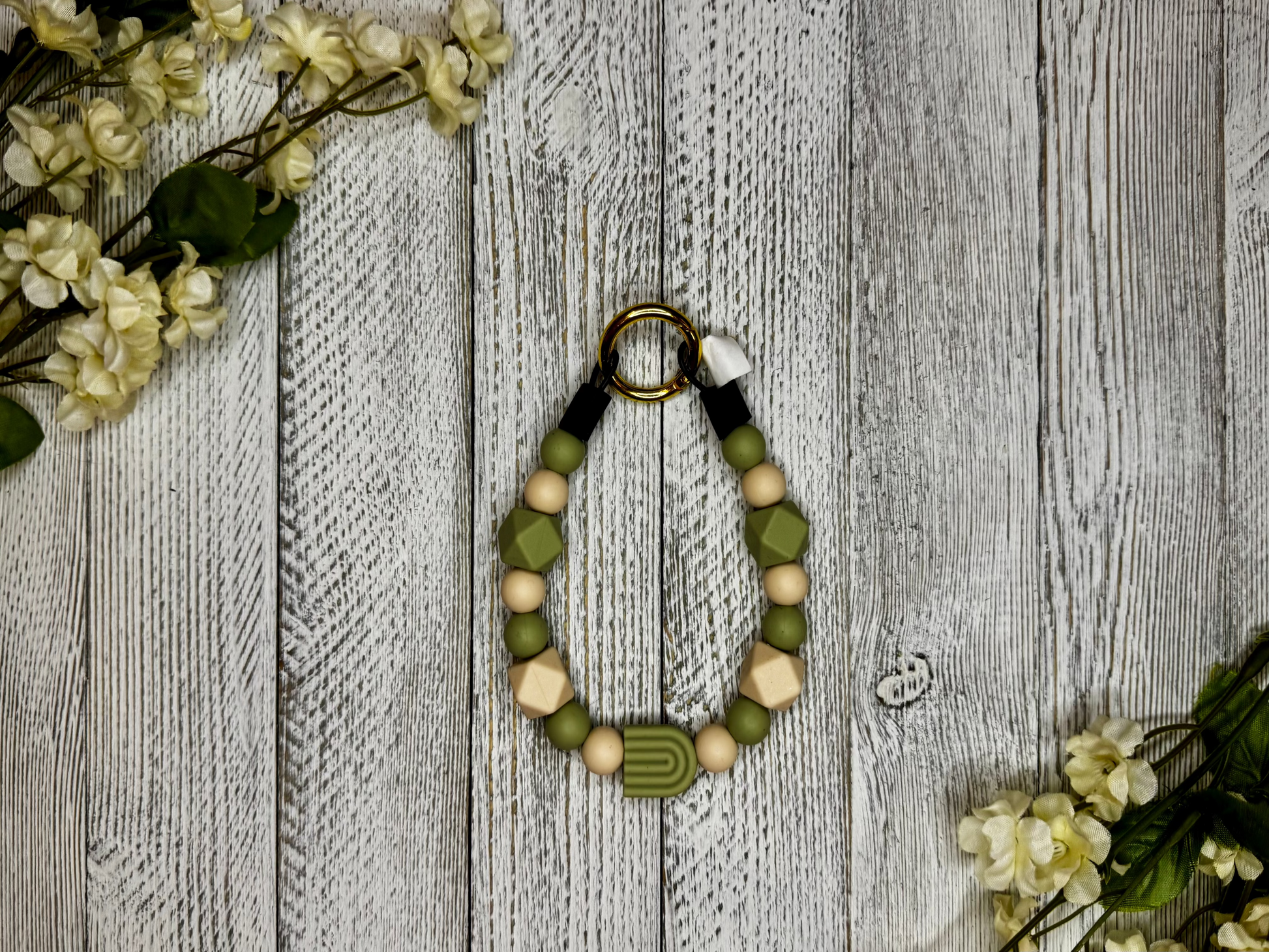 Beaded pet necklace with a olive green and tan pattern, including a rainbow. Displayed on a wooden surface with flowers for decoration. 